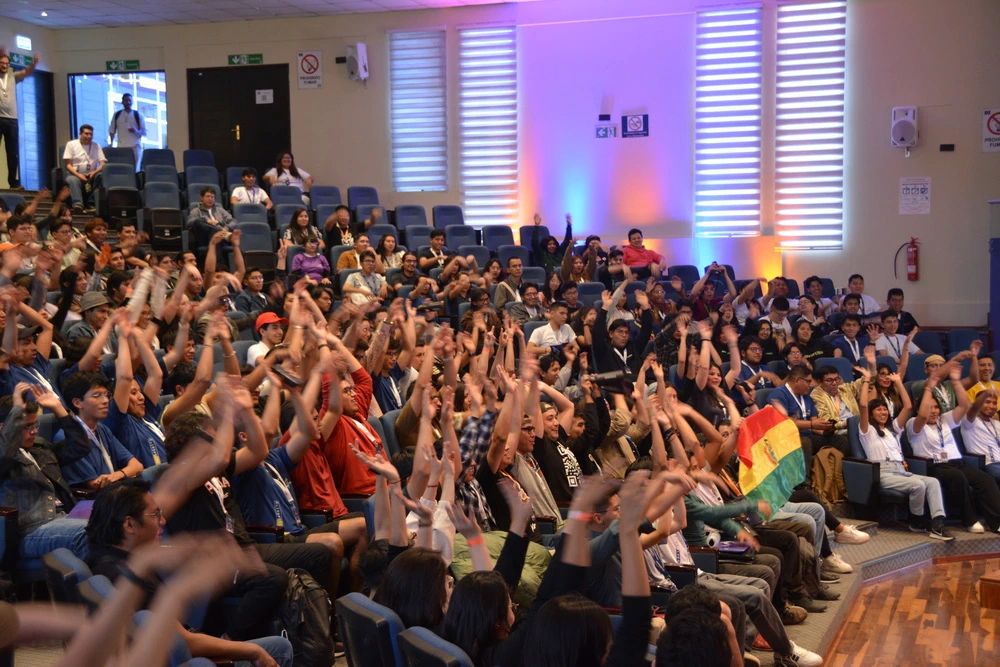 Auditorio lleno en AWS Community Day Bolivia, con asistentes levantando las manos y una bandera boliviana entre el público